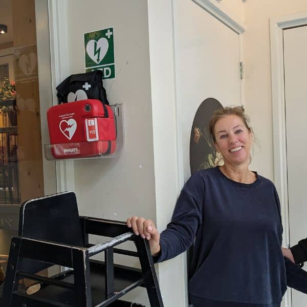 Woman standing next to a wall-mounted defibrillator beside chairs in a waiting area.