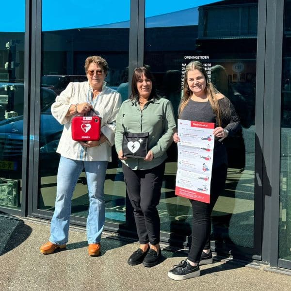 Three people posing outside a building holding a red AED and an instructional board.
