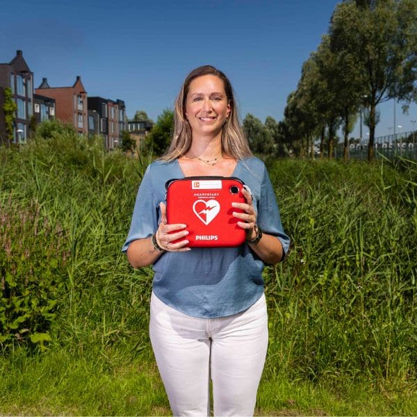 Woman standing in a field holding a red defibrillator, smiling at the camera.